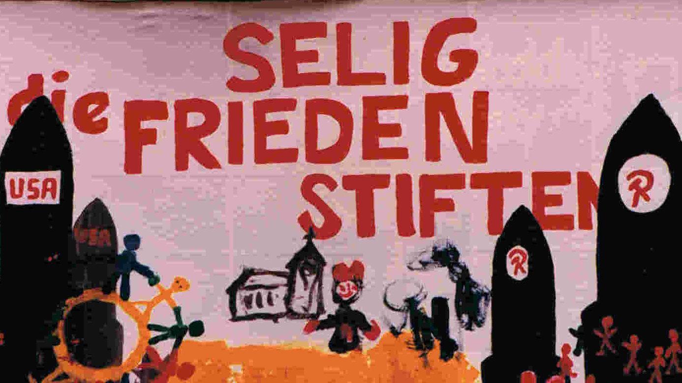 Auf einem selbstgemalten Plakat sind schwarze Raketen recht uns links zu sehen. In der Mitte des Bildes befindet sich eine Kirche, davor Menschen. In Rot ist geschrieben: Selig die Frieden stiften.