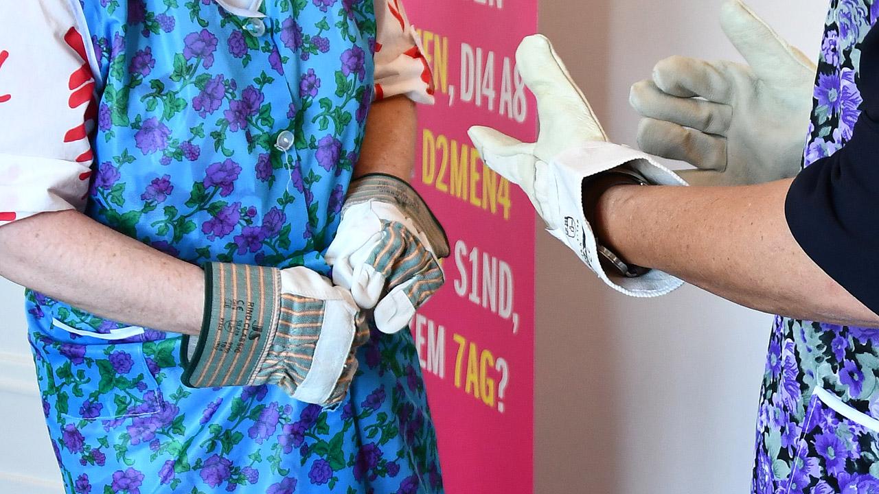 Handmotorik Ausschnitt von Frauen in Kittelschürzen, die mit Arbeitshandschuhen die Knöpfe schließen wollen