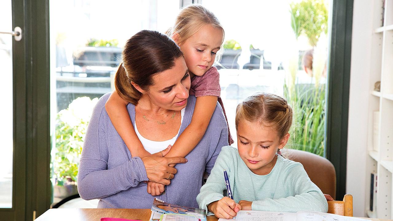 Hilfe bei Hausaufgaben Eine Mutter hilft ihrer Tochter bei den Hausaufgaben, eine andere hat ihre Arme um ihren Hals geschlungen