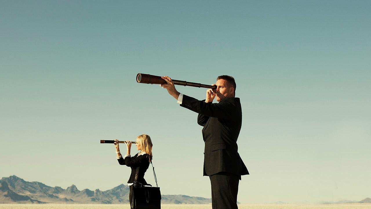 Eine Frau und ein Mann in Business-Outfit mit Teleskop in einer Wüste