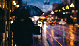 „Eine Person mit Regenschirm steht bei Regen an einer beleuchteten Straße in der Dämmerung. Das unscharfe Stadtlicht erzeugt eine nachdenkliche und ruhige Atmosphäre.“