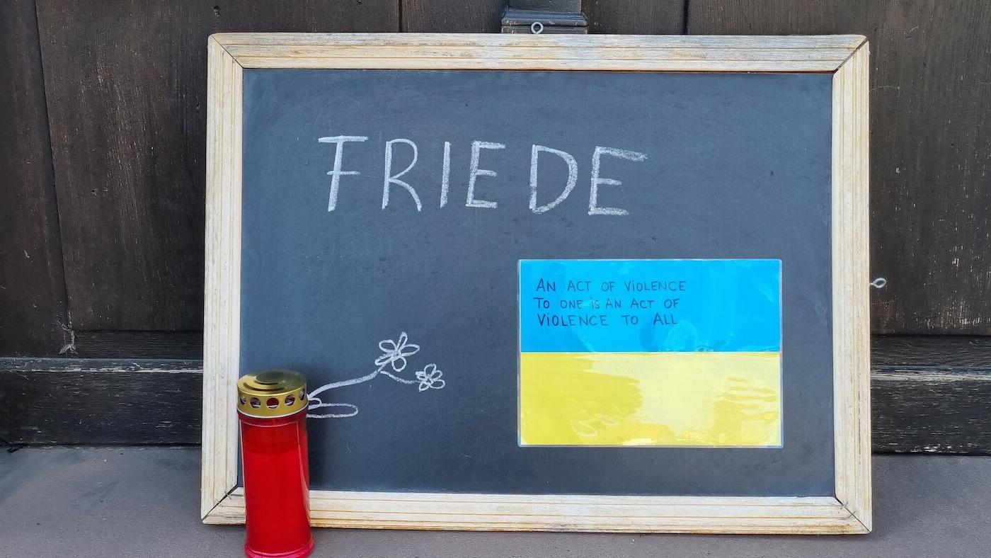 Man sieht eine Kreidetafel vor einer Tür mit der Aufschrift "Friede", davor eine Grabkerze und an der Kreidetafel befestigt ein Schild in den ukrainischen Farben mit dem Text: An Act of Violence to one is an act of violence to all