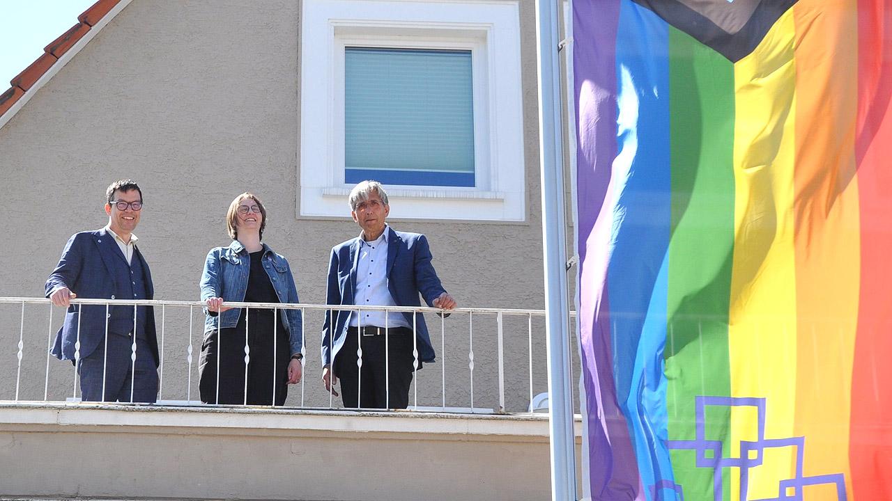Queer Flagge Drei Menschen auf einem Balkon, daneben eine Flagge mit Regenbogenfarben, oben dem Transgenderzeichen und EKHN-Logo