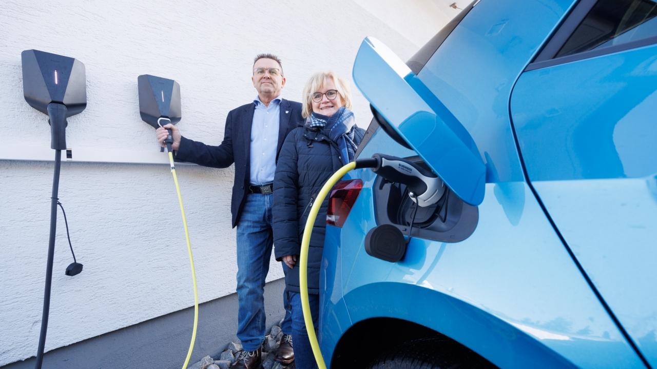 Ein Mann und eine Frau stehen neben einem blauen Elektroauto, das über eine Wallbox an der Hauswand geladen wird. Beide halten Ladekabel in der Hand und blicken in die Kamera.