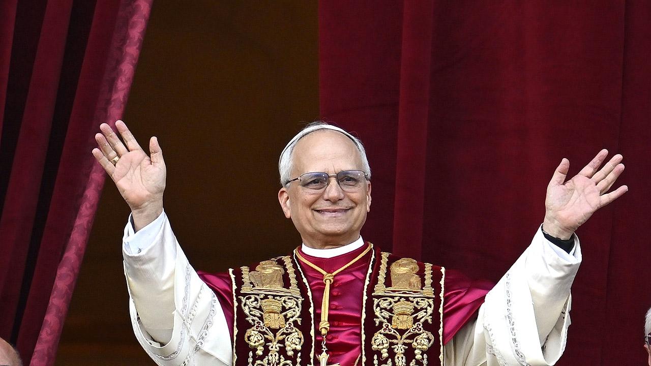 Papst Leo XIV. hebt die Hände und trägt ein weißes Gewand, darüber ein kürzeres bordeauxfarbene Seidencape und eine schwarz-goldene Stola um den Hals