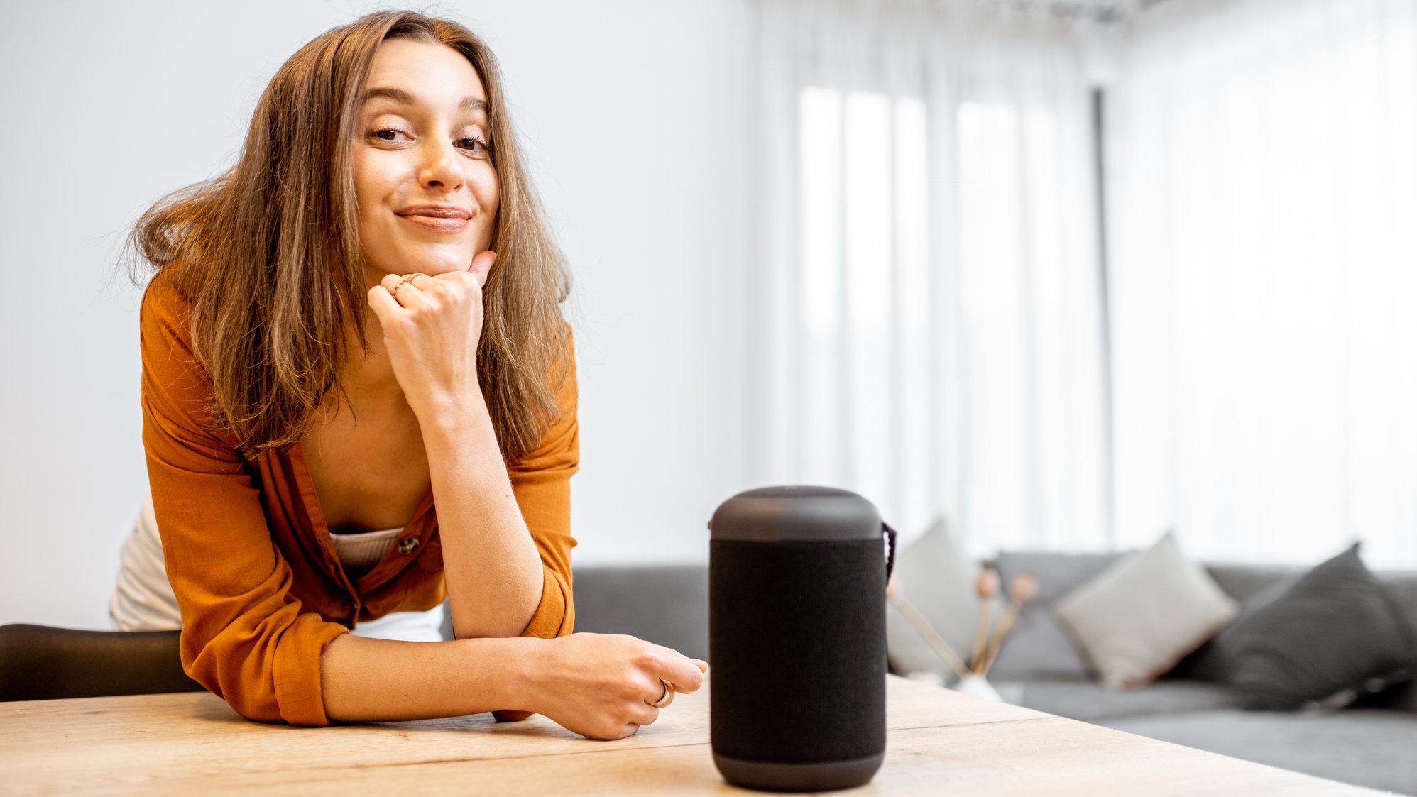 Eine Frau schaut lächelnd in die Kamera, sie hat ihre Arme auf den Tisch gestützt vor ihr steht die Sprachassistentin Alexa