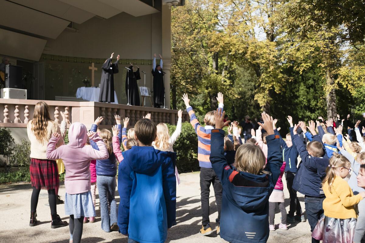 Segen_Tauffest Bad Homburg Gottesdienstbesucherinnen - vorwiegend Kinder - von hinten aufgenommenstehen mit erhobenen Armen und blicken zur Bühne mit dem Altar. Die Veranstaltung findet in einem Park mit Bäumen statt. Auf der Bühne 3 Pfarrer:innen ebenfalls mit erhobenen Armen.