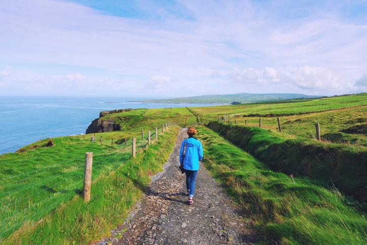 Segen_Irland_Irischer_Reisesegen_Freundschaft_Wegbegleitung Eine Frau mit blauer Jacke wandert einen Pfad am Meer entlang zwischen grünen Wiesen.