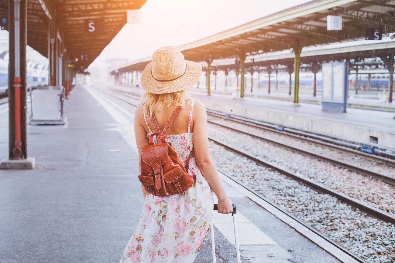 Frau steht mit einem Sommerkleid und Koffer und Rucksack auf dem Bahnhof am Gleis  und schaut in die Ferne.