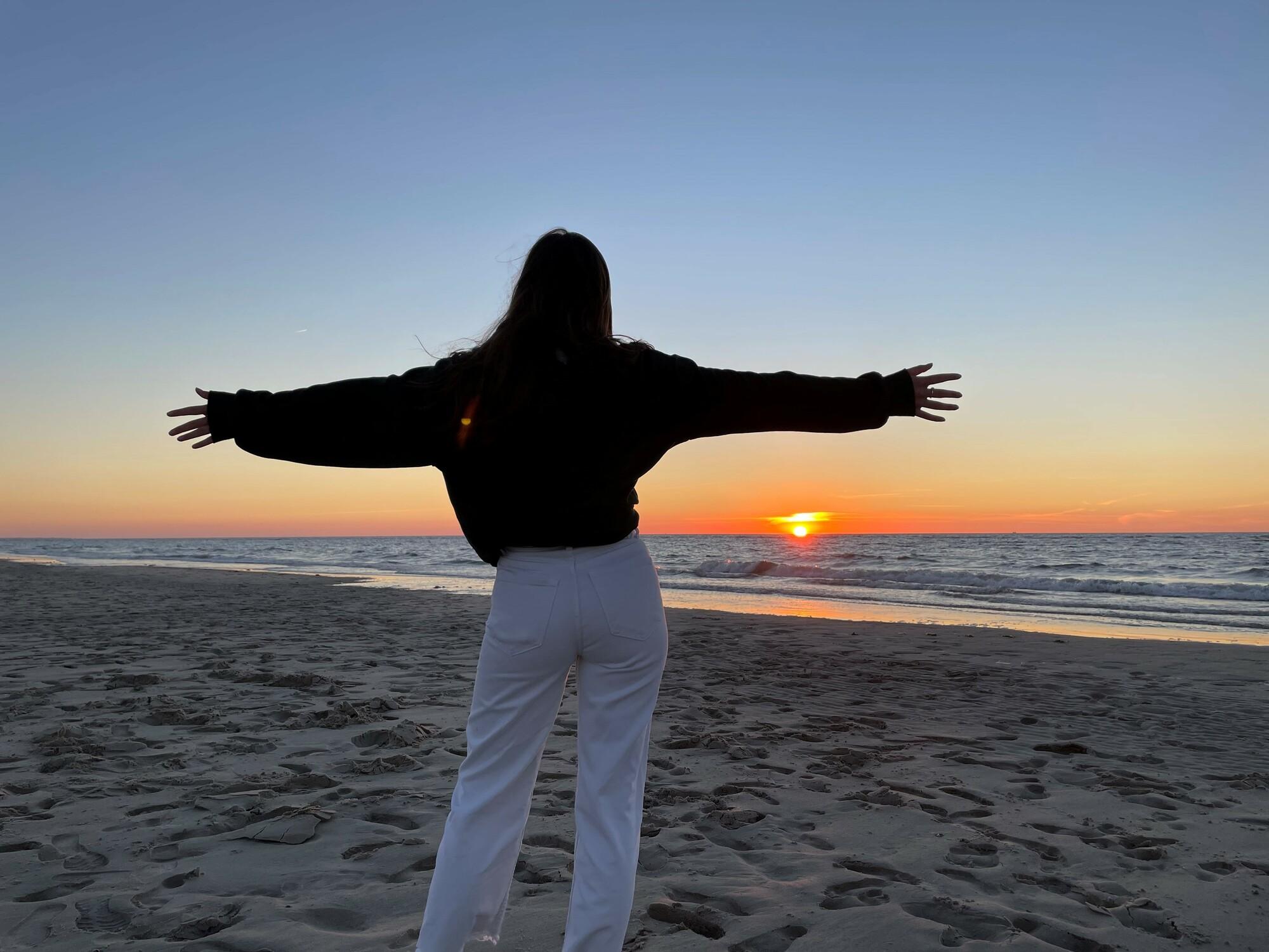 Eine Frau in weißer Hose und schwarzer Jacke steht am Strand und schaut auf das Meer. Die Sonne geht gerade unter, der Himmel färbt sich rot und gold. Sie hält die Arme ausgebreitet, zum Segen bereit.