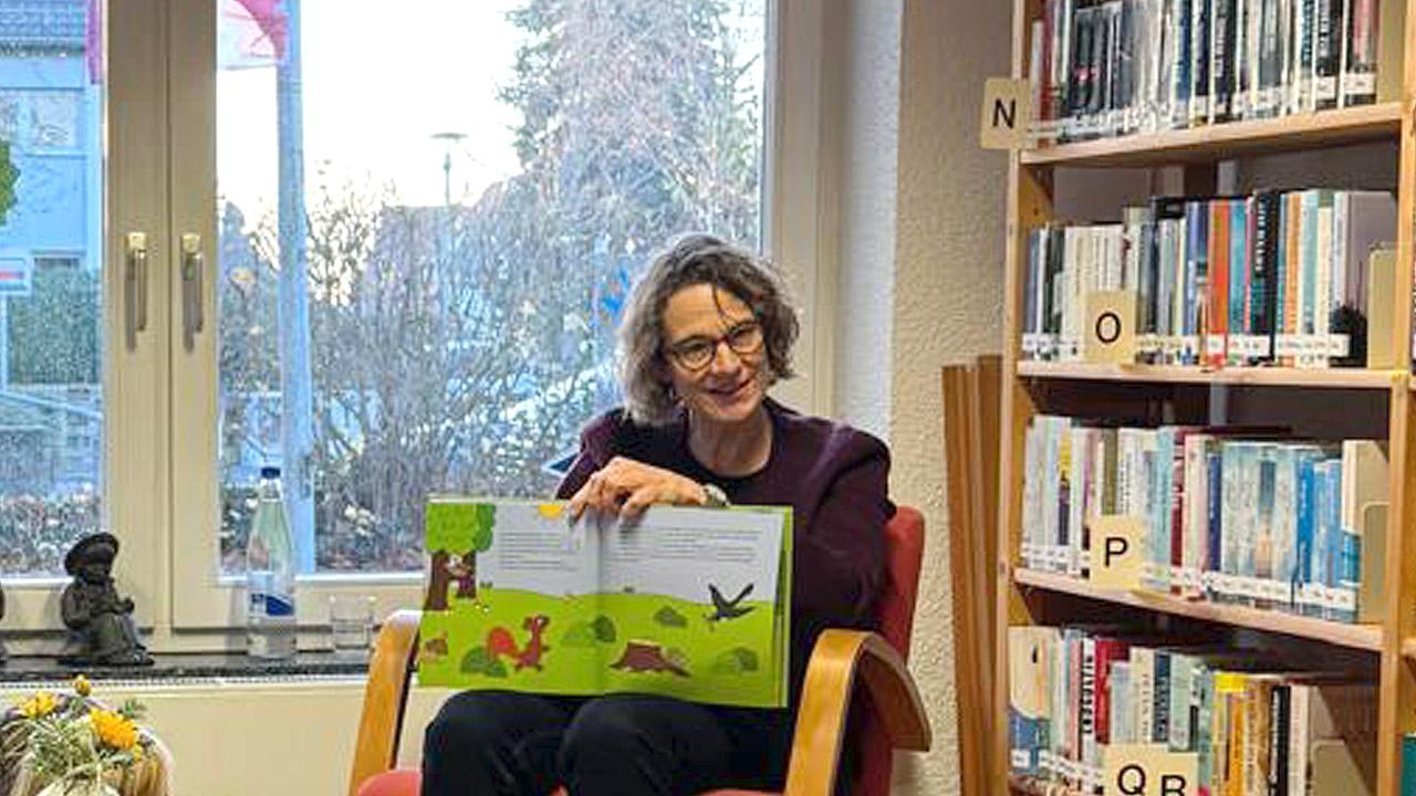 Die Kirchenpräsidentin sitzt mit einem aufgeschlagenen Kinderbuch auf einem Stuhl