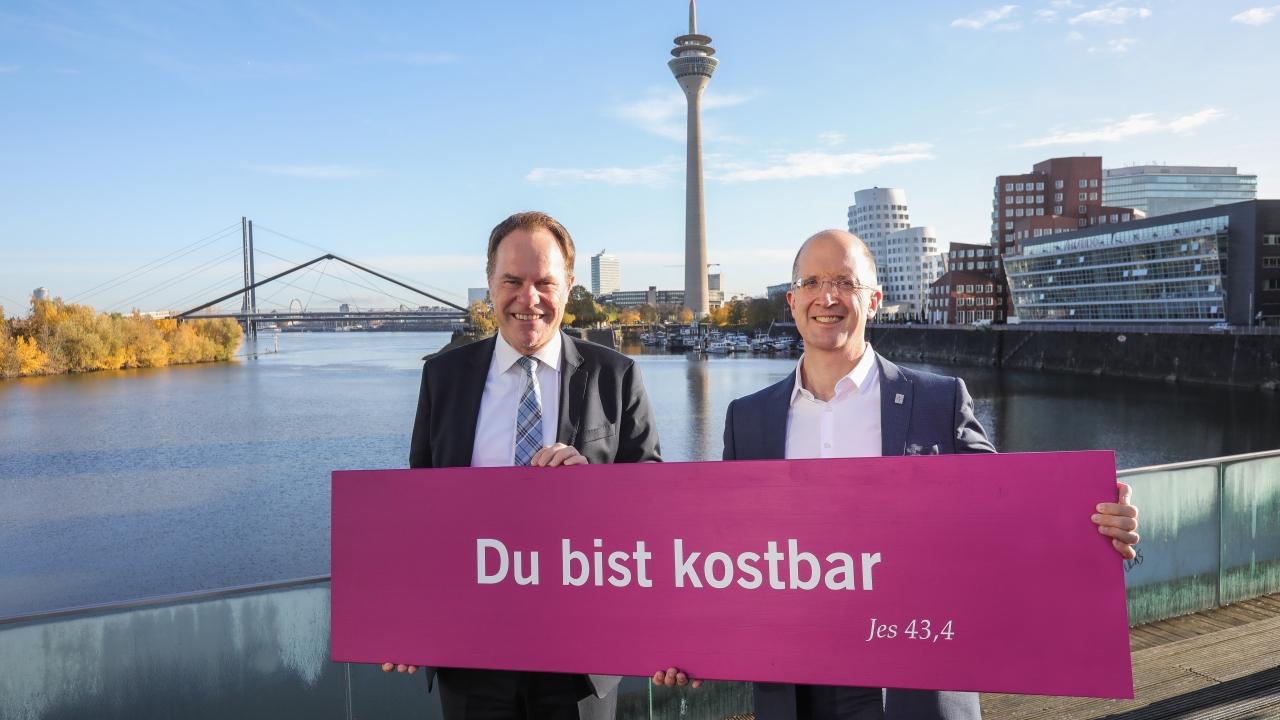 Zwei Männer in Anzügen stehen am Rheinufer vor der Düsseldorfer Skyline mit Rheinturm im Hintergrund und halten ein pinkes Schild mit der Aufschrift „Du bist kostbar (Jes 43,4)“.