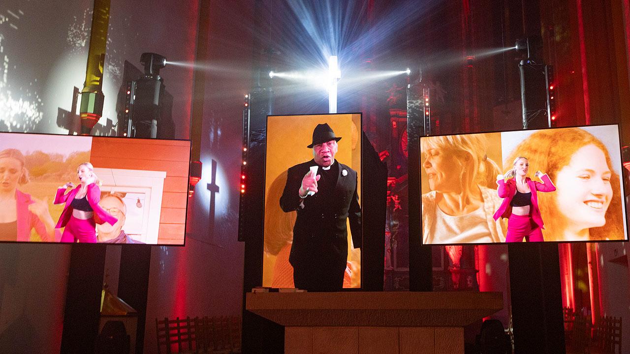 Erlebnis:Kirche  Ein singender Mann mit Hut in der Mitte, daneben Bildschirme mit einer Tänzerin
