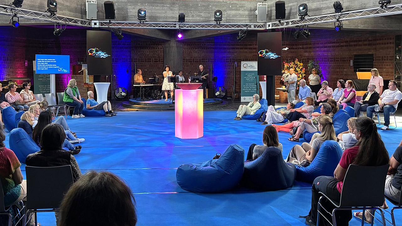 Quellenkirche Menschen auf Bänken und Loungesesseln rund um den rötlich beleuchteten Pult mit der kleinen Wasserfontaine