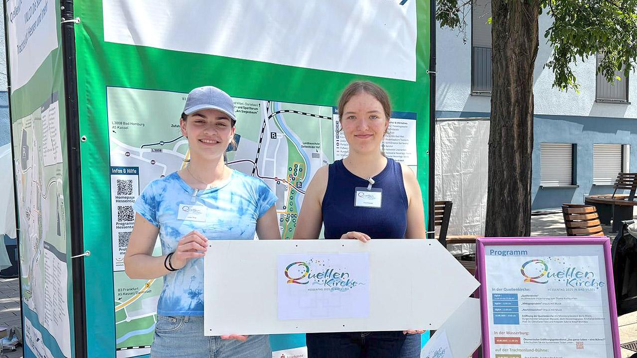 Zwei junge Frauen halten ein Schild mit der Aufschrift "Quellenkirche"