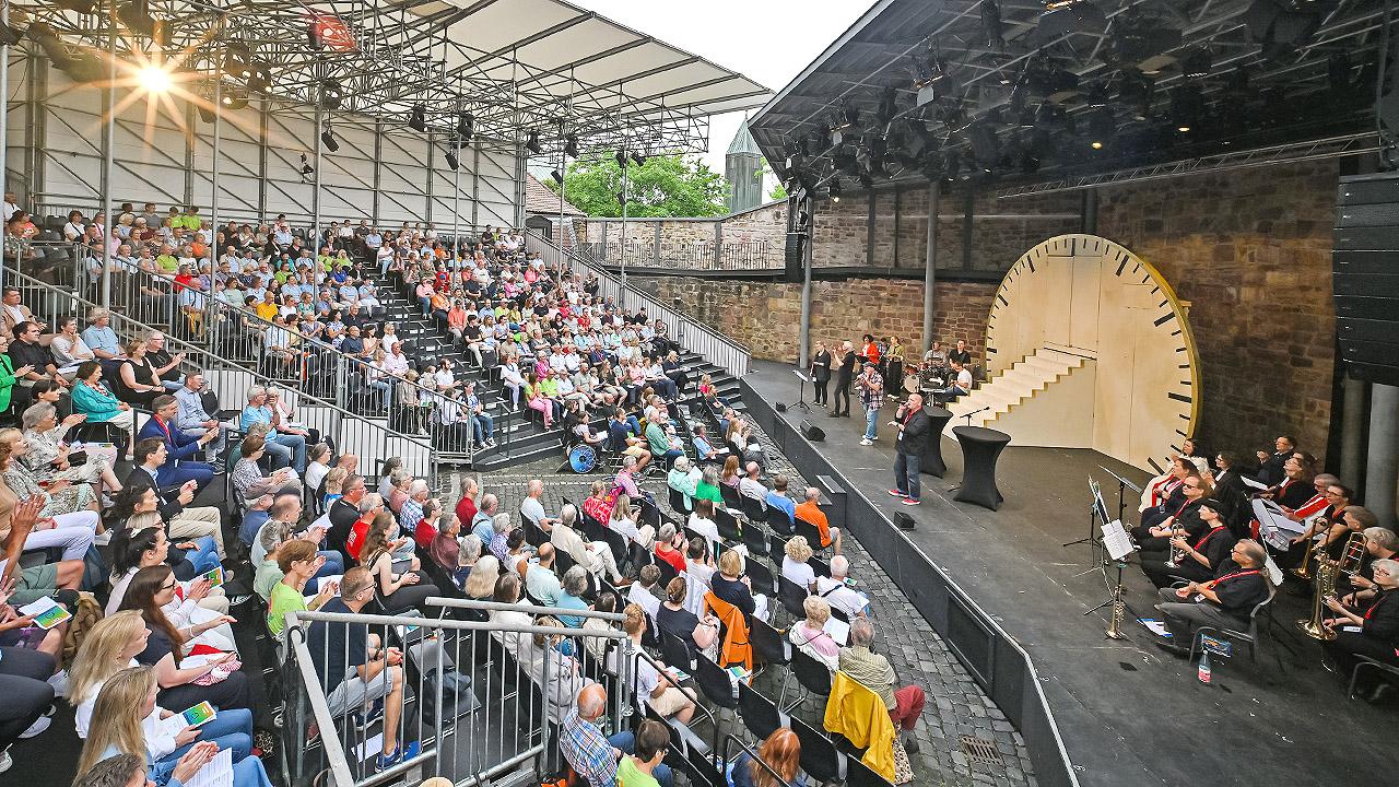 Ökumenischer Gottesdienst Viele Menschen in einer Art Outdoor-Theater, unten eine Bühne mit überdimensionaler Uhr