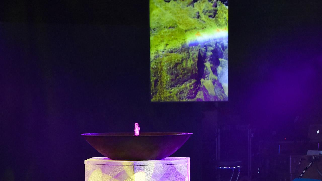 Multimedia Schale mit kleiner Wasserfontaine, dahinter ein Lichtbild aus der grünen Natur
