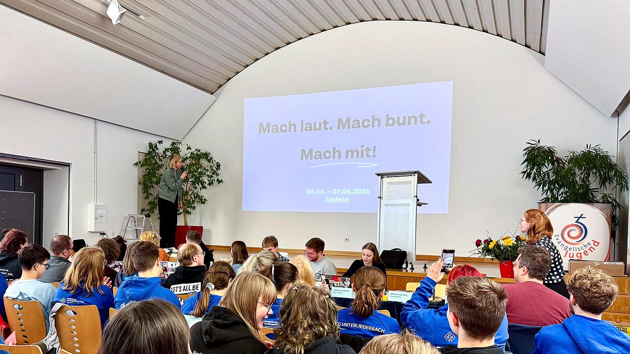EJHN Publikum vor einem Rednerpult, dahinter wird das Motto mit einem Beamer an die Wand geworfen: „Mach laut. Mach bunt. Mach mit!“