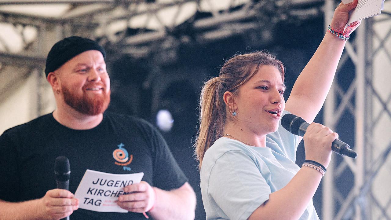 Moderierende Ein junger Mann mit Bart un deine junge Frau mit Mikrophon, die Frau hebt einen Arm voller Energie nach oben