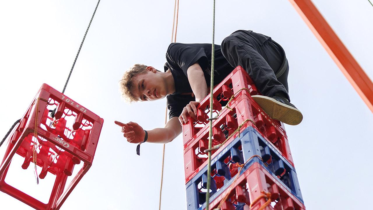 Kistenklettern Junge kniet oben auf einem Turm voller Getränkekisten und nimmt die nächste Kiste zum Stapeln in Empfang