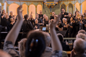 Aus dem Publikum heraus fotografiert mit vordergründiger Unschärfe auf einen Gospelchor, in schwarz gekleidtet in einem Altarraum stehend. Viele klatschende Hände